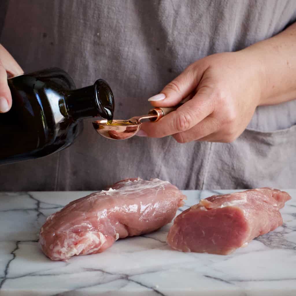 Womans hands pouring olive oil from a dark brown bottle into a gold tablespoon over a piece of uncooked pork