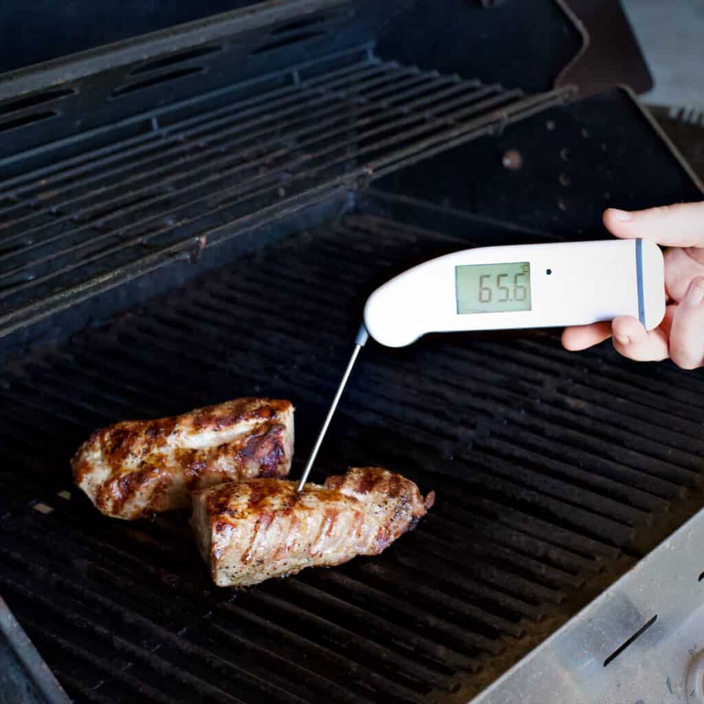 White digital thermometer taking the temperature of two pieces of pork cooking on a gas BBQ