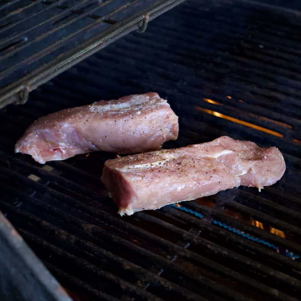 Two pieces of pork fillet cooking on a the black metal bars of a gas BBQ