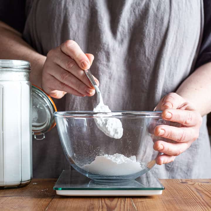 womans hands spooning icing sugar into a glass bowl on a wooden counter