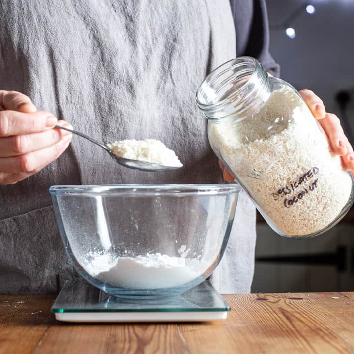 womans hands holding a glass jar of desiccated coconut and a silver spoon of coconut over a glass bowl