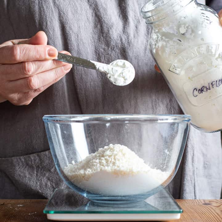 womans hand tipping cornflour from a silver spoon into a glass mixing bowl