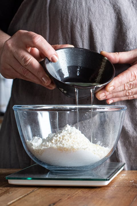 womans hands pouring an egg white from a small black bowl into a larger glass bowl filled with white baking ingredients