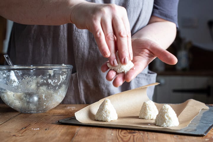 womans hands moulding a little coconut pyramid cake over the top of a baking tray filled with more coconut cakes