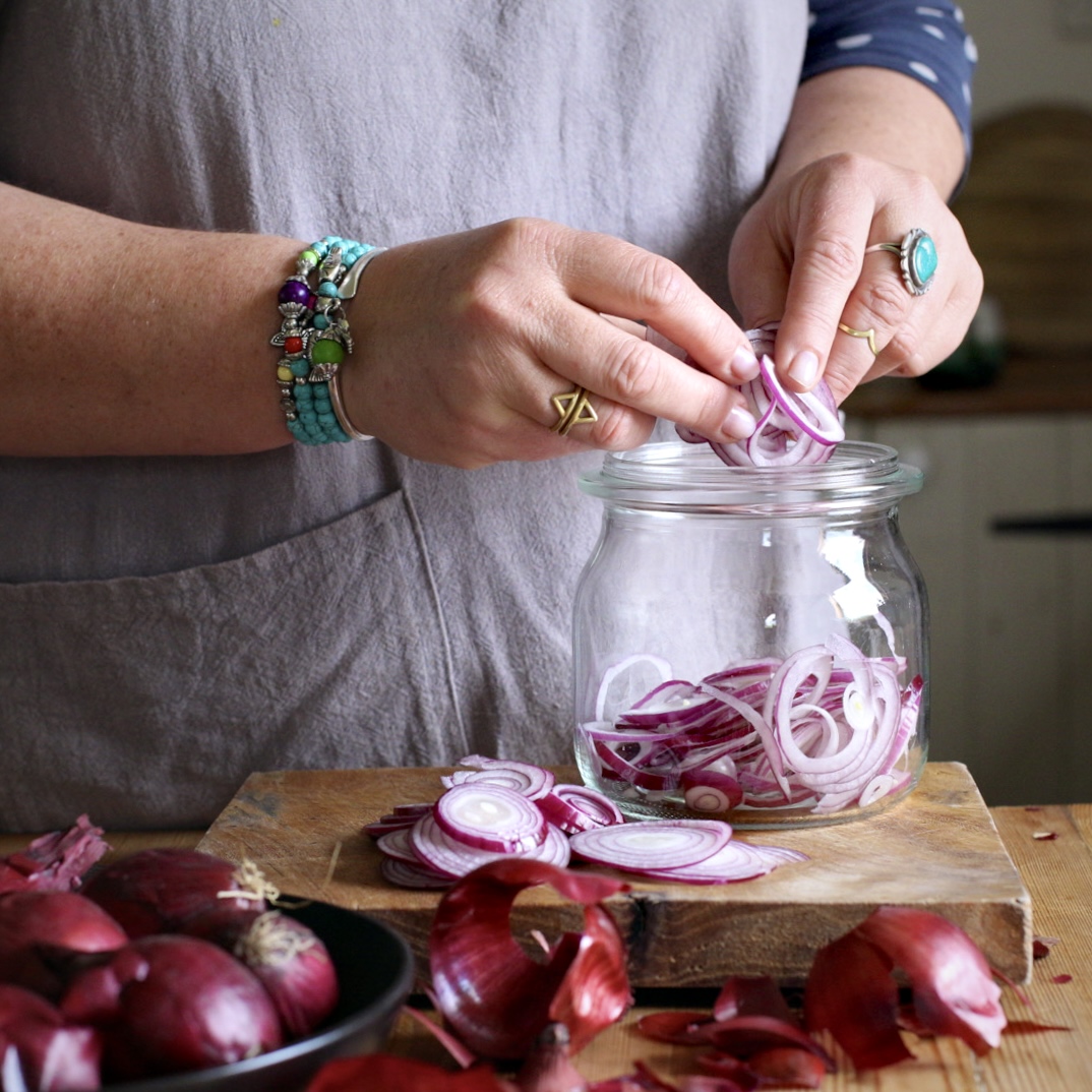 Womans hands with turquoise jewellery on placing rings of sliced red onions into a glass jar on a wooden kitchen counter