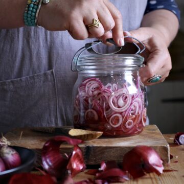Womans in grey sealing an old fashioned glass jar filled with pickled red onions