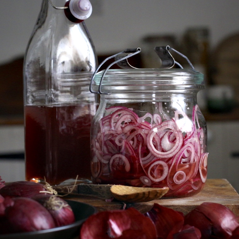 Old fashioned glass jar of pickled red onions next to a glass bottle of red wine vinegar and several red onions skins