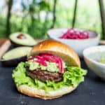 garden table scene with burger in brioche bun, mashed avocado and pickled red onions and various ingredients in background