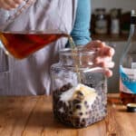 woman in grey pouring brandy from a glass jug into a big glass jar of blackcurrants