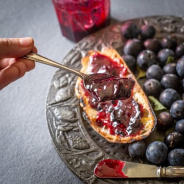 a spoon of wild sloe and blackberry jam held over a piece of toast and damsons on a pewter plate
