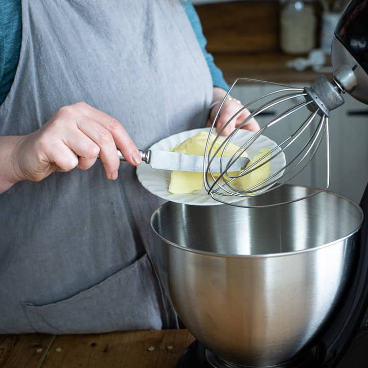 womans hands scraping lumps of butter from a white plate into a stainless steel mixing bowl