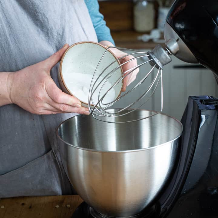 womans hands tipping white sugar from a small bowl into a silver mixing bowl