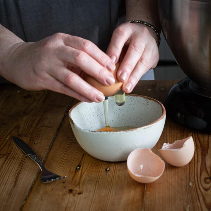 womans hands cracking eggs into a small white bowl on a wooden kitchen counter