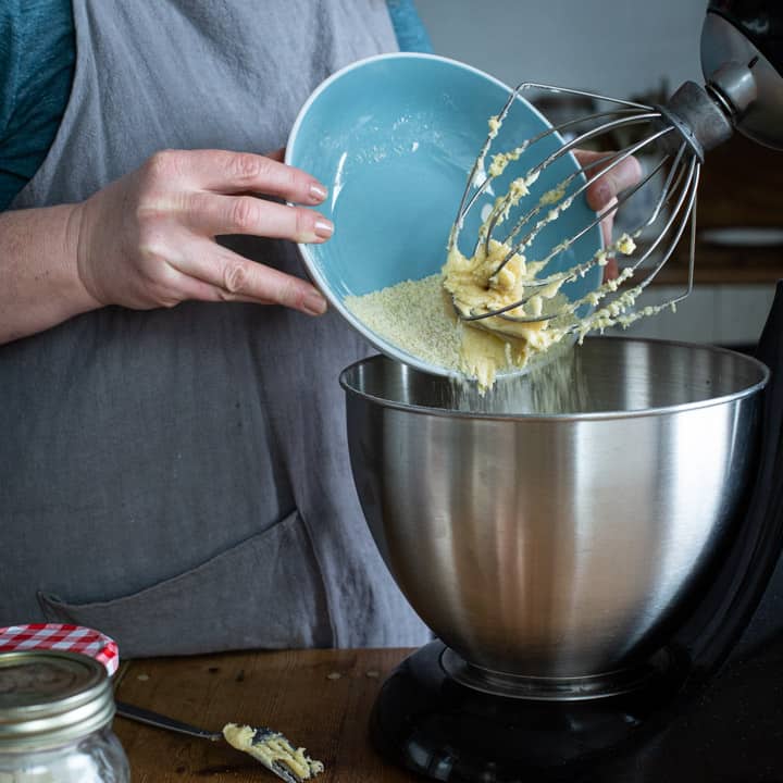 woman tipping ground almonds from a blue bowl into a silver electric mixer