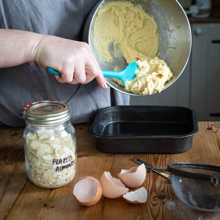 woman scraping yellow cake batter from a silver bowl into a black baking tin