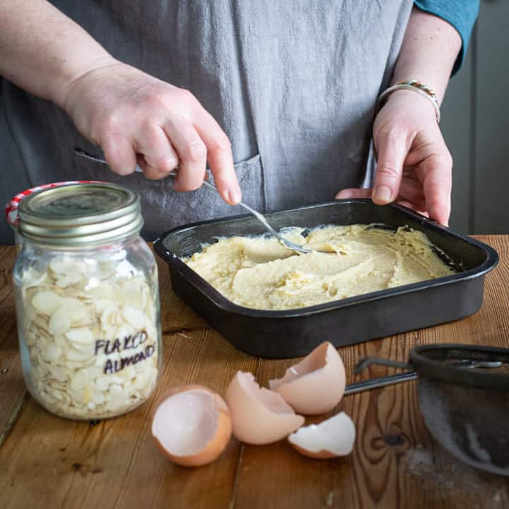 womans hands smoothing out almond batter in black square cake tin