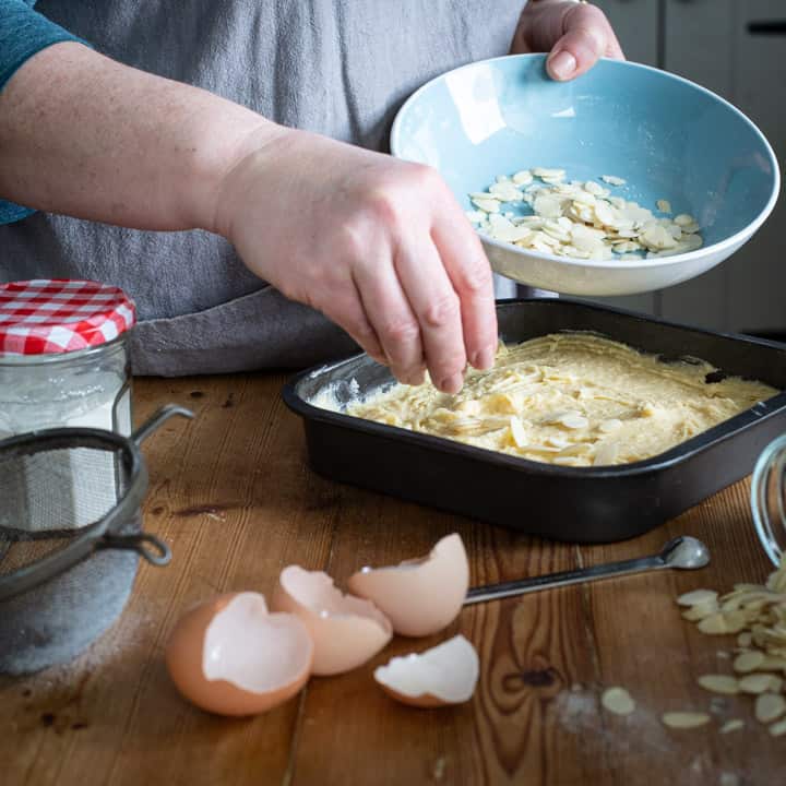 womans hands scattering flaked almonds over a black baking tin filled with cake batter