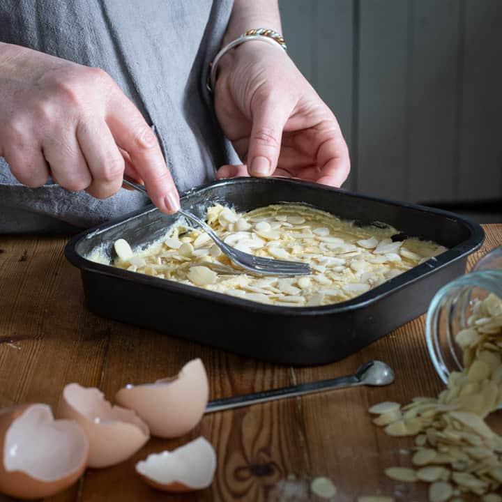 womans hands smoothing out almond batter in black square cake tin