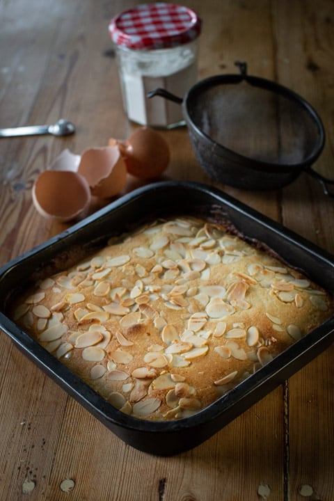 black square baking tin filled with golden brown almond cake surrounded by broken eggs shells and baking mess