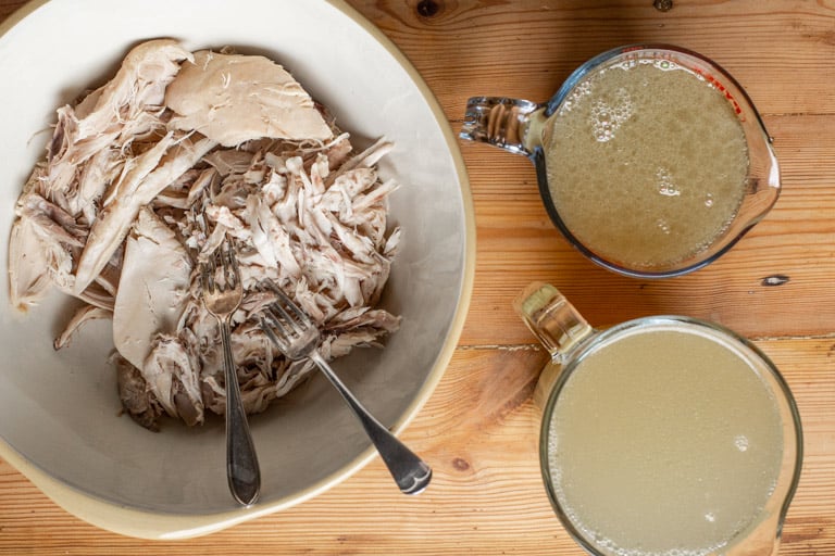 wooden background with shredded slow cooker whole chicken and 2 jugs of homemade chicken stock