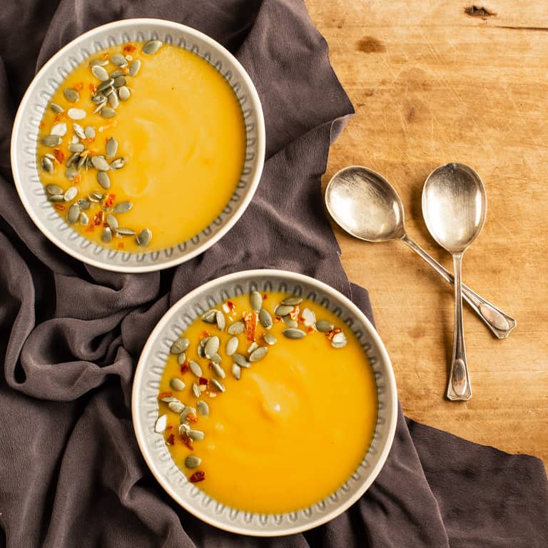 wooden background with two bowl of dairy free pumpkin soup sprinkled with pumpkin seeds and chilli flakes