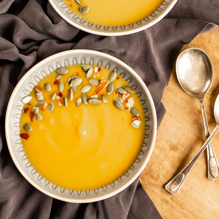 wooden background with two bowl of dairy free pumpkin soup sprinkled with pumpkin seeds and chilli flakes