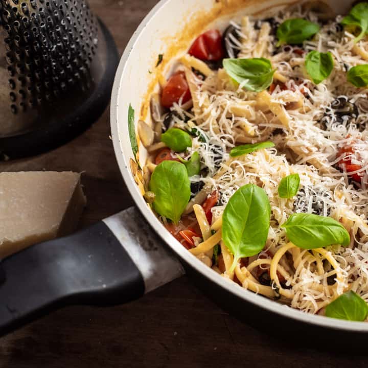 wooden background with black and white saute pan filled with one pot veggie pasta, fresh parmesan and basil leaves