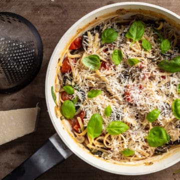 wooden background with black and white saute pan filled with one pot veggie pasta, fresh parmesan and basil leaves