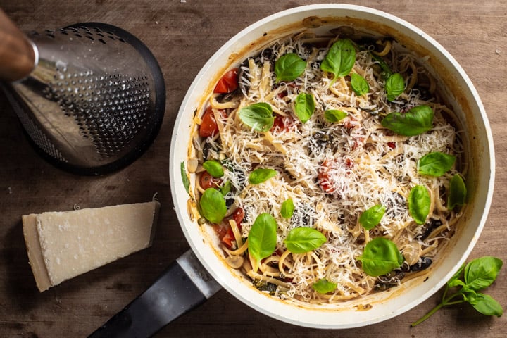 wooden background with black and white saute pan filled with one pot veggie pasta, fresh parmesan and basil leaves