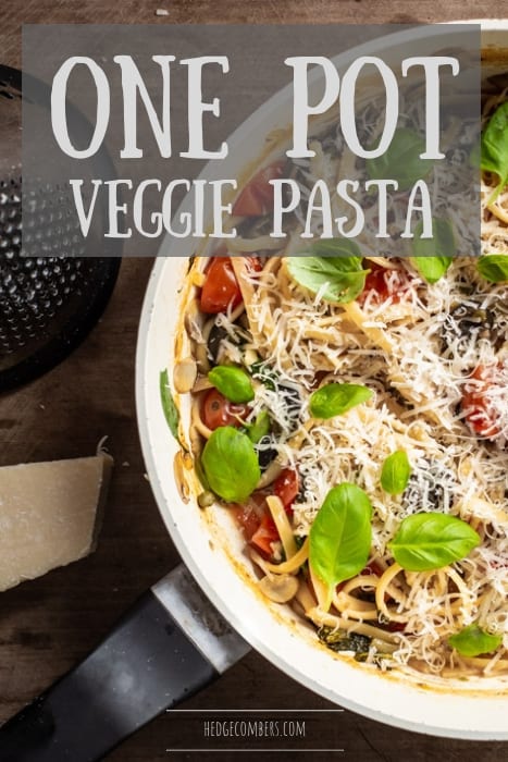 wooden background with black and white saute pan filled with one pot veggie pasta, fresh parmesan and basil leaves