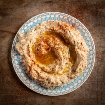 wooden bakground with pretty plate filled with homemade hummus drizzled with olive oil and smoked paprika