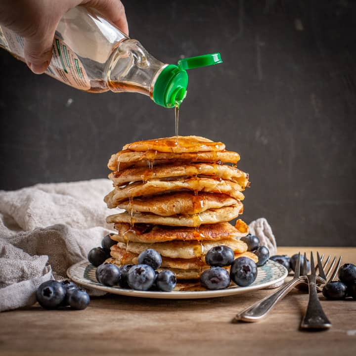 black background with stack of eggless pancakes with blueberries and maple syrup being poured over