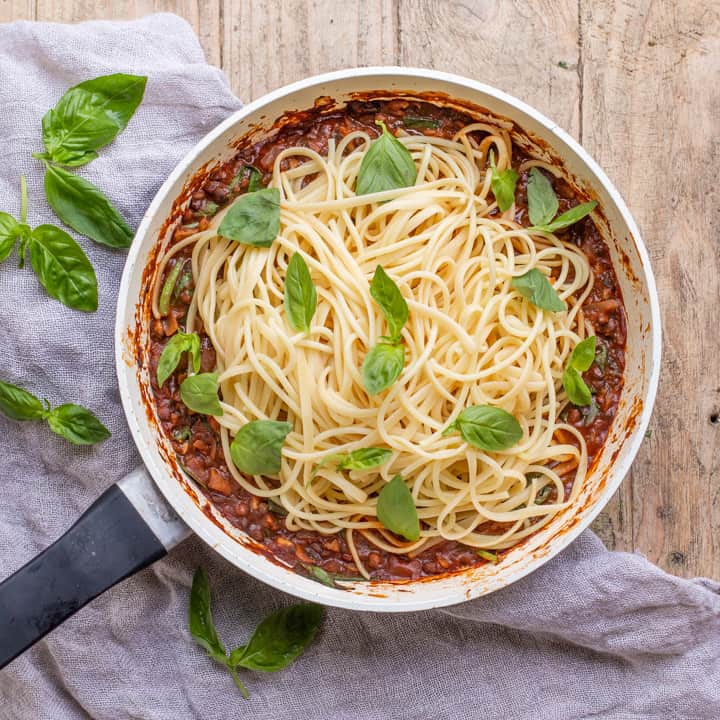 wooden background with white pan with vegan spaghetti bolognese piled up and scattered with fresh basil leaves