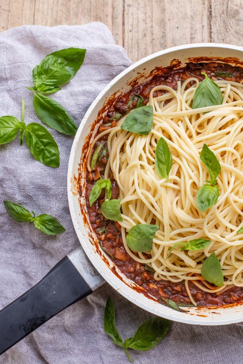 wooden background with white pan with vegan spaghetti bolognese piled up and scattered with fresh basil leaves