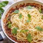 wooden background with white pan with vegan spaghetti bolognese piled up and scattered with fresh basil leaves