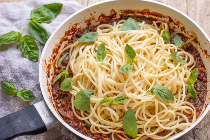 wooden background with white pan with vegan spaghetti bolognese piled up and scattered with fresh basil leaves