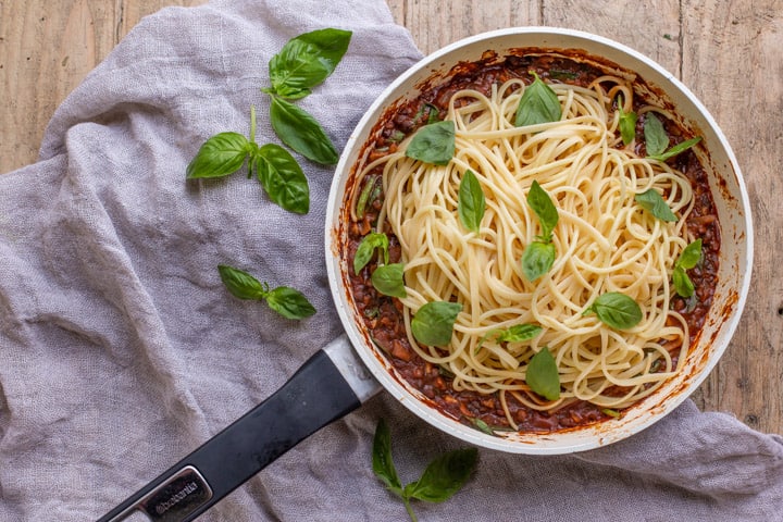 wooden background with white pan with vegan spaghetti bolognese piled up and scattered with fresh basil leaves