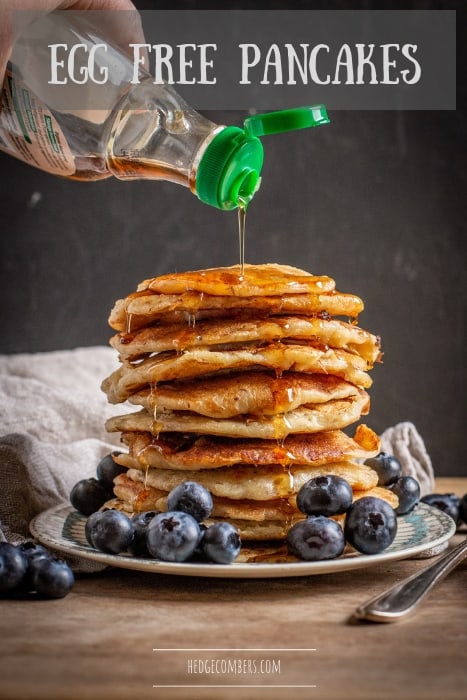 black background with stack of eggless pancakes with blueberries and maple syrup being poured over
