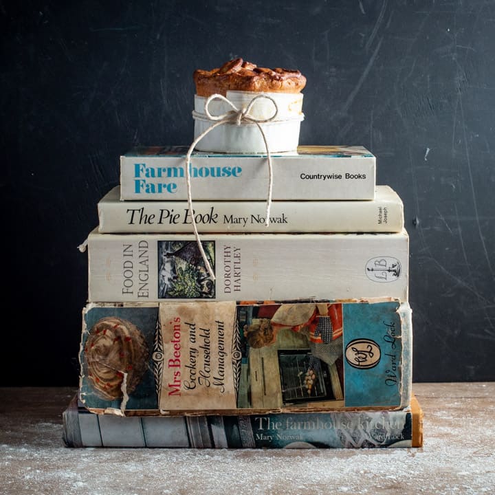 black backdrop with a pile of vintage cookbooks and a single homemade British pork pie balanced on top