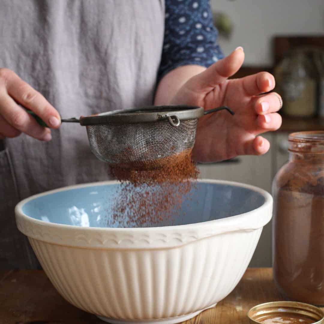 Woman in grey sifting cocoa powder into a blue and white mixing bowl