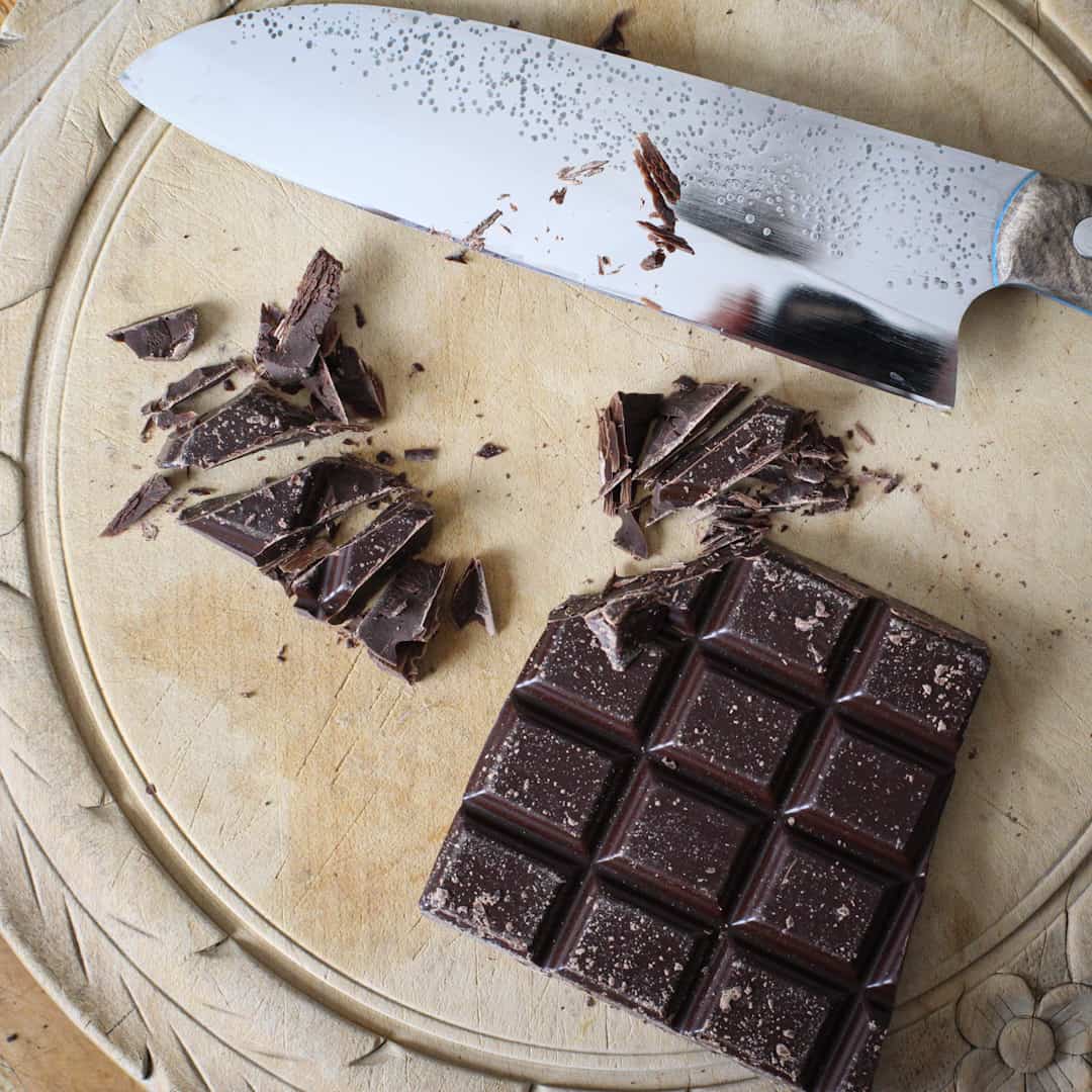 Wooden chopping board with a bar of dark chocolate getting chopped into small pieces with a silver knife