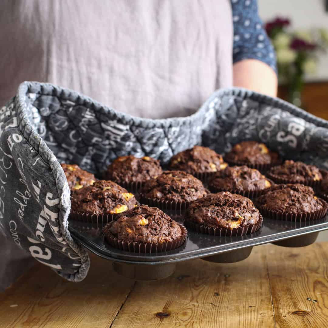Woman with black oven gloves on holding a black tray of freshly baked banana chocolate muffins
