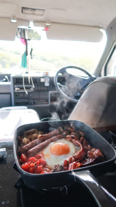 cast iron breakfast skillet being made in a camper van with views of the drivers seat, sterring wheel and green fields beyond