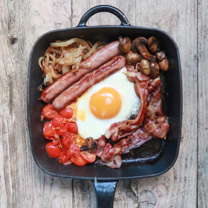 wooden background with cast iron breakfast skillet filled with breakfast foods like sausages, bacon, eggs, mushrooms etc