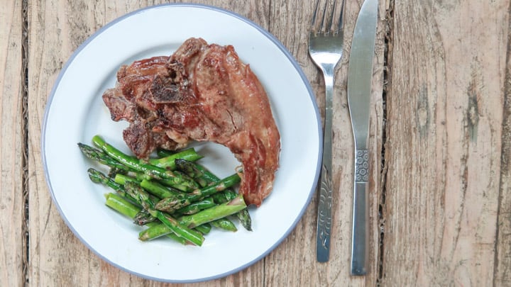 wooden background with white plate and cutlery, with 2 lamb chops and cooked asparagus