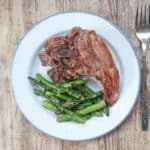 wooden background with white plate and cutlery, with 2 lamb chops and cooked asparagus