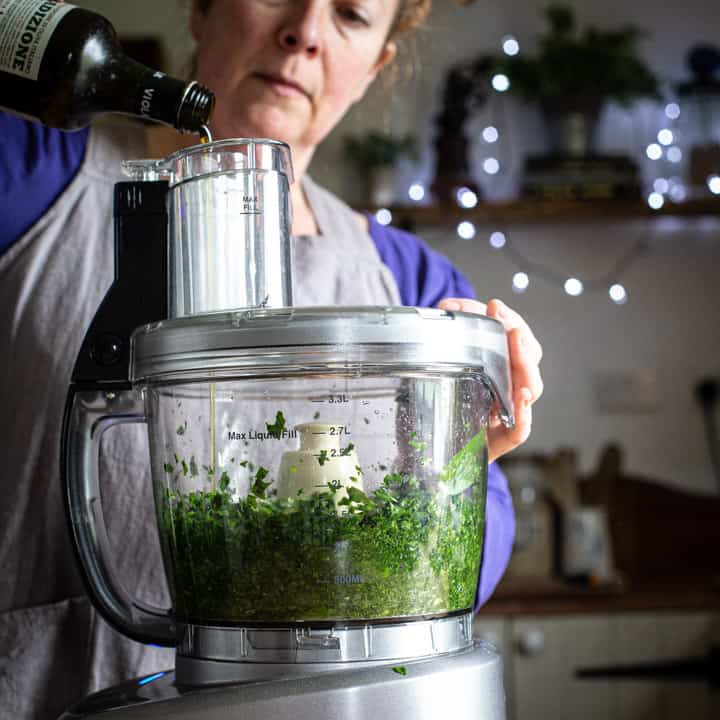 woman in grey pouring olive oil into a food processor to make fresh pesto