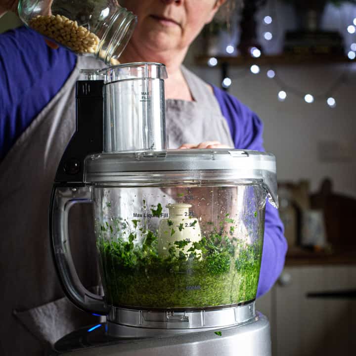 woman in grey pouring nuts into a food processor to make pesto