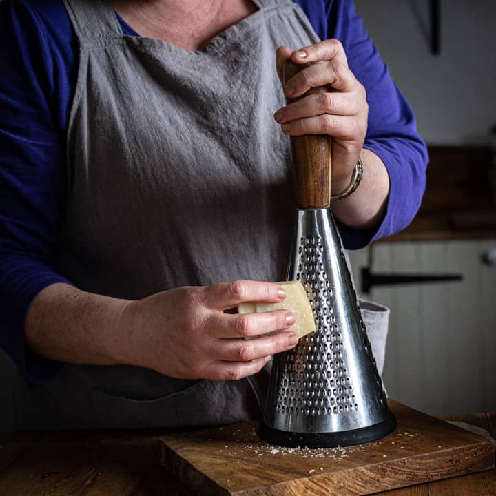 woman in grey grating a block of parmesan cheese onto a wooden board