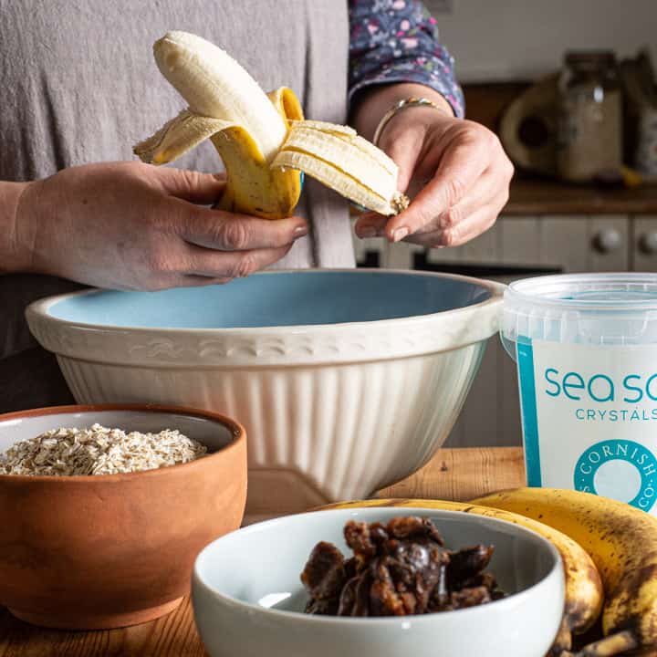 woman in grey peeling a banana in a kitchen scene surrounded with baking ingredients